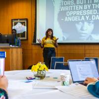 woman in yellow at the front of a room, with a quote from Angela Davis behind her